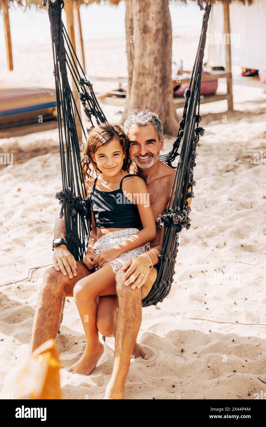 Porträt eines lächelnden Vaters und einer Tochter, die auf einer Hängematte am Strand sitzen Stockfoto