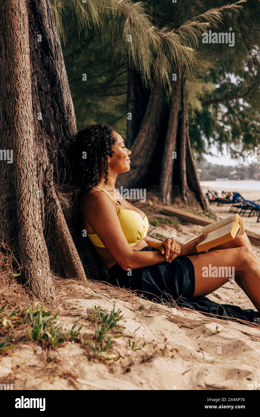 Seitenansicht einer reiferen Frau, die auf einem Baumstamm mit Buch am Strand liegt Stockfoto