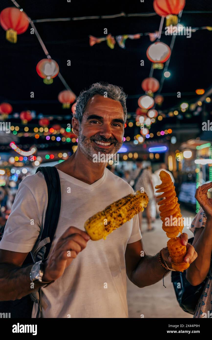 Porträt eines lächelnden Mannes, der Tornado-Kartoffeln in der Hand hält, gebratene und gegrillte Mais auf dem Lebensmittelmarkt im Urlaub Stockfoto