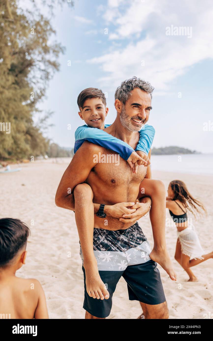 Ein lächelnder Mann ohne Hemd, der einen Huckepack hat, als er am Strand spaziert Stockfoto