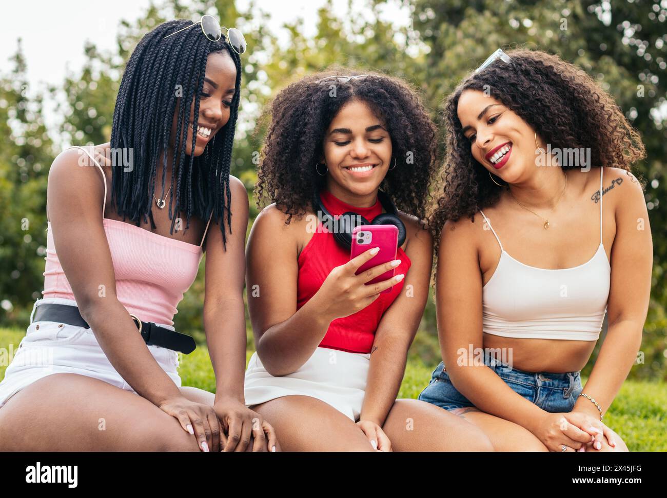 Drei afroamerikanische Mädchen sitzen in einem Park und haben Spaß. Sie verwenden ein Smartphone und sehen Bilder und Videos aus sozialen Netzwerken Stockfoto