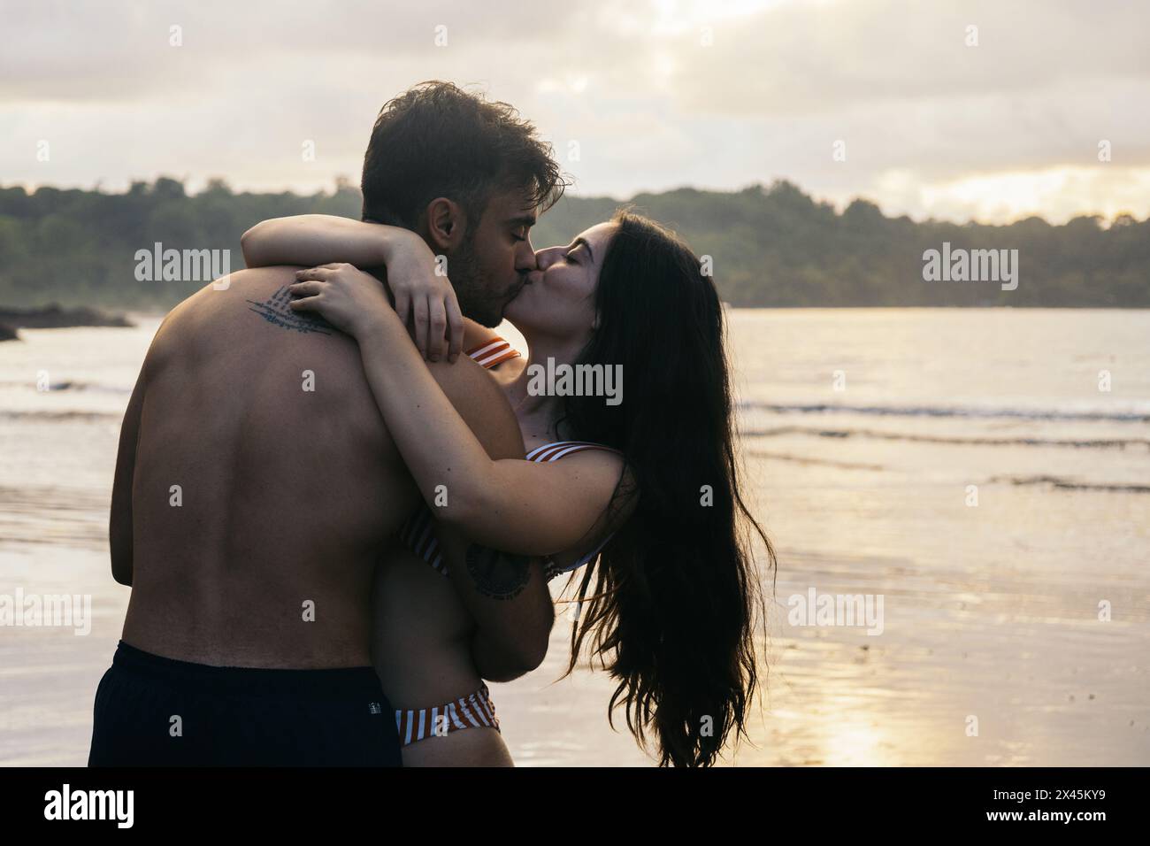 Romantisches Foto von einem Paar, das sich am Strand küsst und den Sonnenuntergang beobachtet Stockfoto