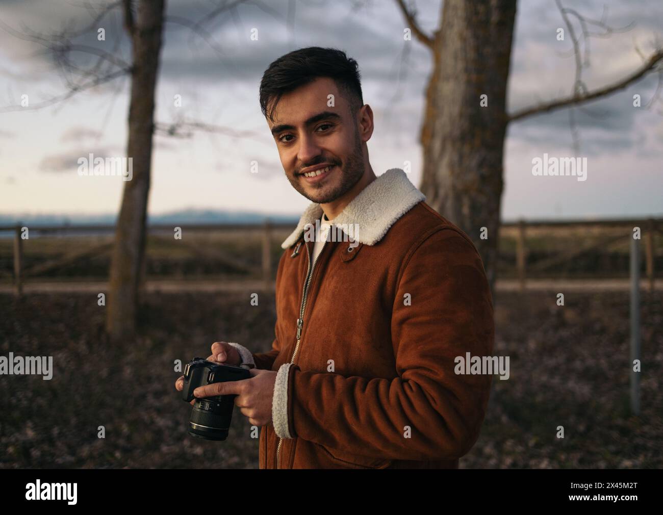 Porträt eines lächelnden Fotografen, der in die Kamera schaut. Junger Mann in brauner Jacke, der sich die Fotos auf der Kamera ansieht Stockfoto