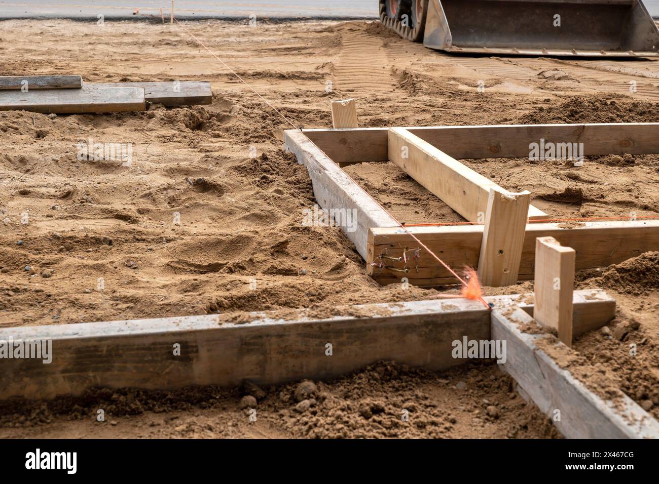 Eine Holzform zur Zementplatzierung im braunen Schmutz auf einer Baustelle. Zwei mal vier Bretter, Schnur, Schmutz und ein Frontlader. Stockfoto