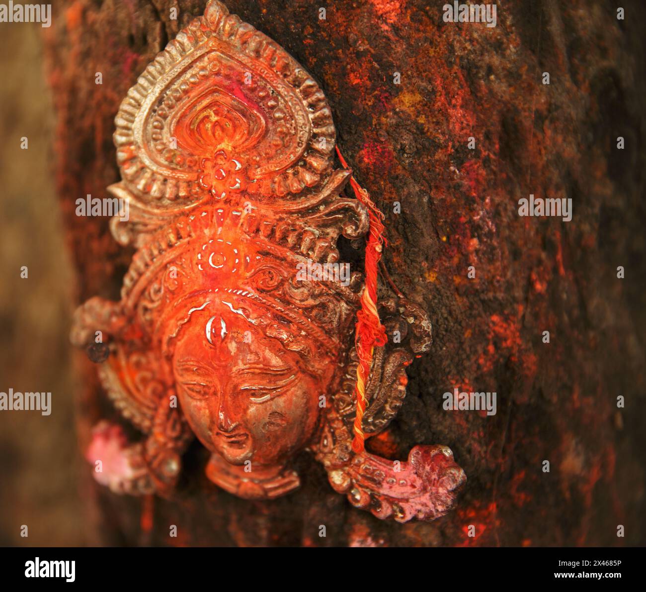 Metallgesicht einer Gottheit, die an einem Baumstamm befestigt ist, bei ASI Ghat in Varanasi, Indien. Stockfoto