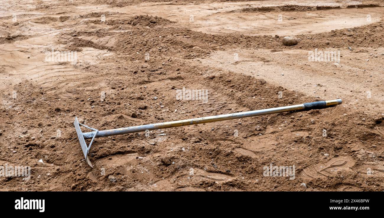 Ein Rechen mit Metallgriff zum Nivellieren von Erde und Schmutz liegt auf dem braunen Boden einer Hausbaustelle. Stockfoto