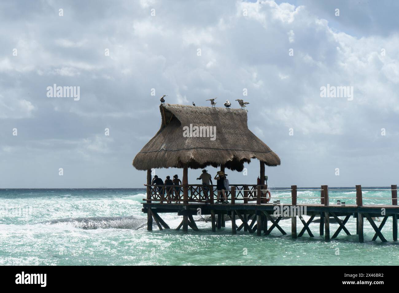 Holzpier mit Strohdach-Hütte auf dem Meer mit Pelikanen ruhend, bewölktem Himmel, Gruppe von Touristen, die vor Regen schützen und den Horizont beobachten, Xcalacoco Me Stockfoto