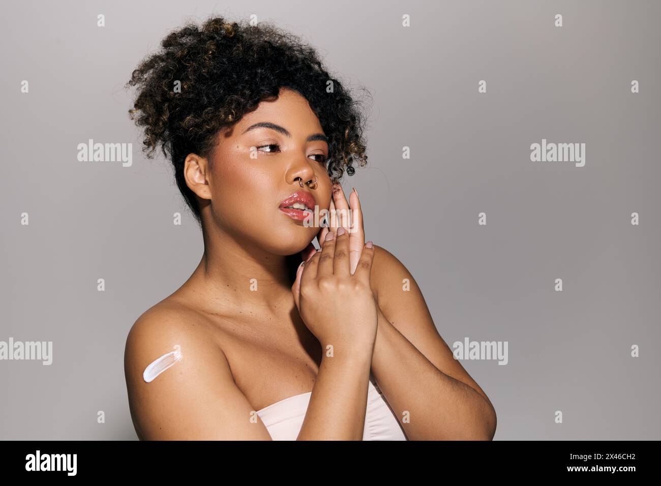 Eine junge afroamerikanische Frau mit lockigen Haaren in einem trägerlosen Oberteil hält ihre Hand im Studio zart an ihr Gesicht. Stockfoto