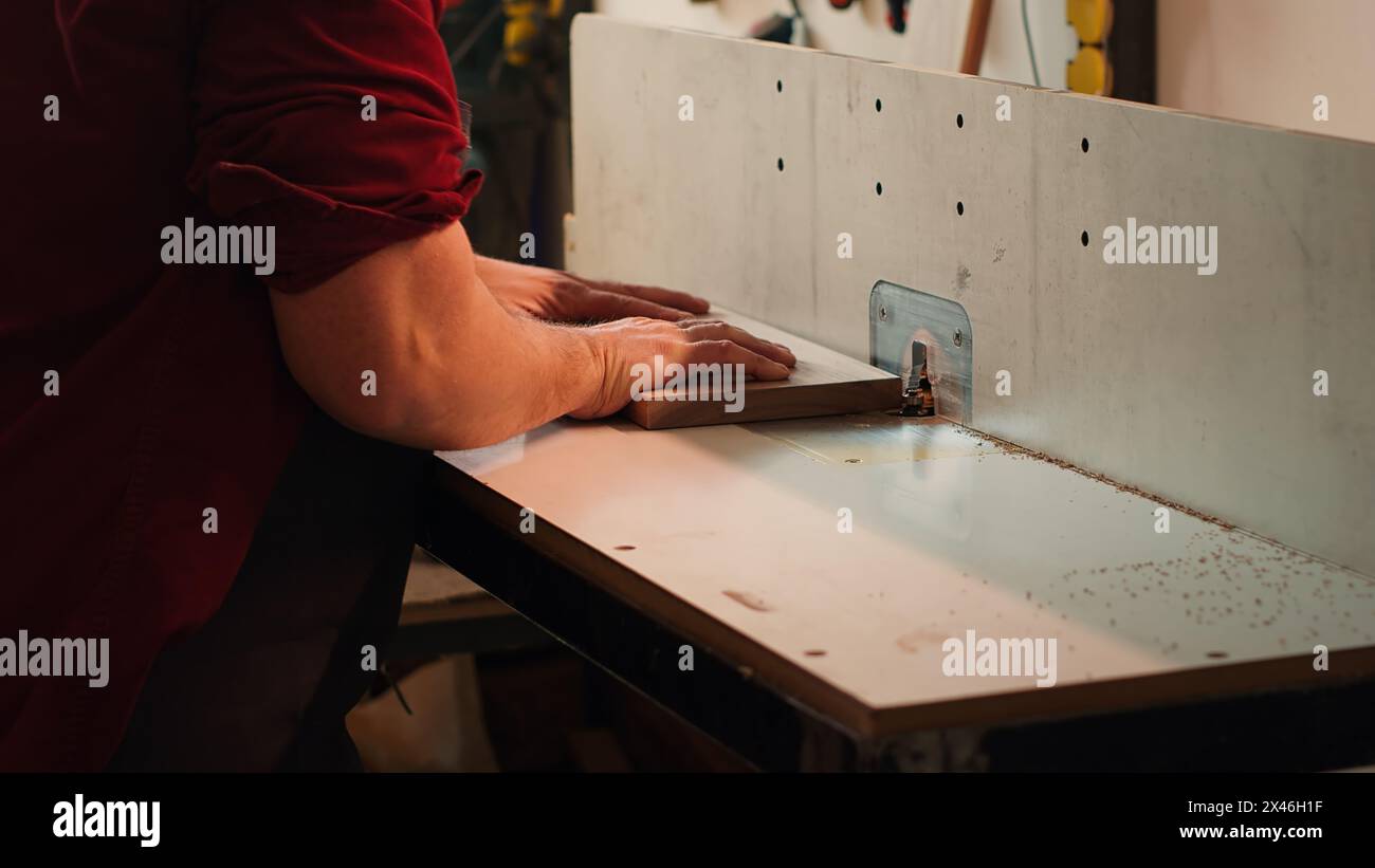 Holzbearbeitungsexperte trägt Schutzkleidung und setzt die Planke in der Spindelmaschine neben dem Kollegen ein. Der CNC-Maschinenbauer verwendet eine Schutzbrille beim Schneiden von Holz mit einem Holzformer, der von seinem Kollegen, Kamera A, unterstützt wird Stockfoto