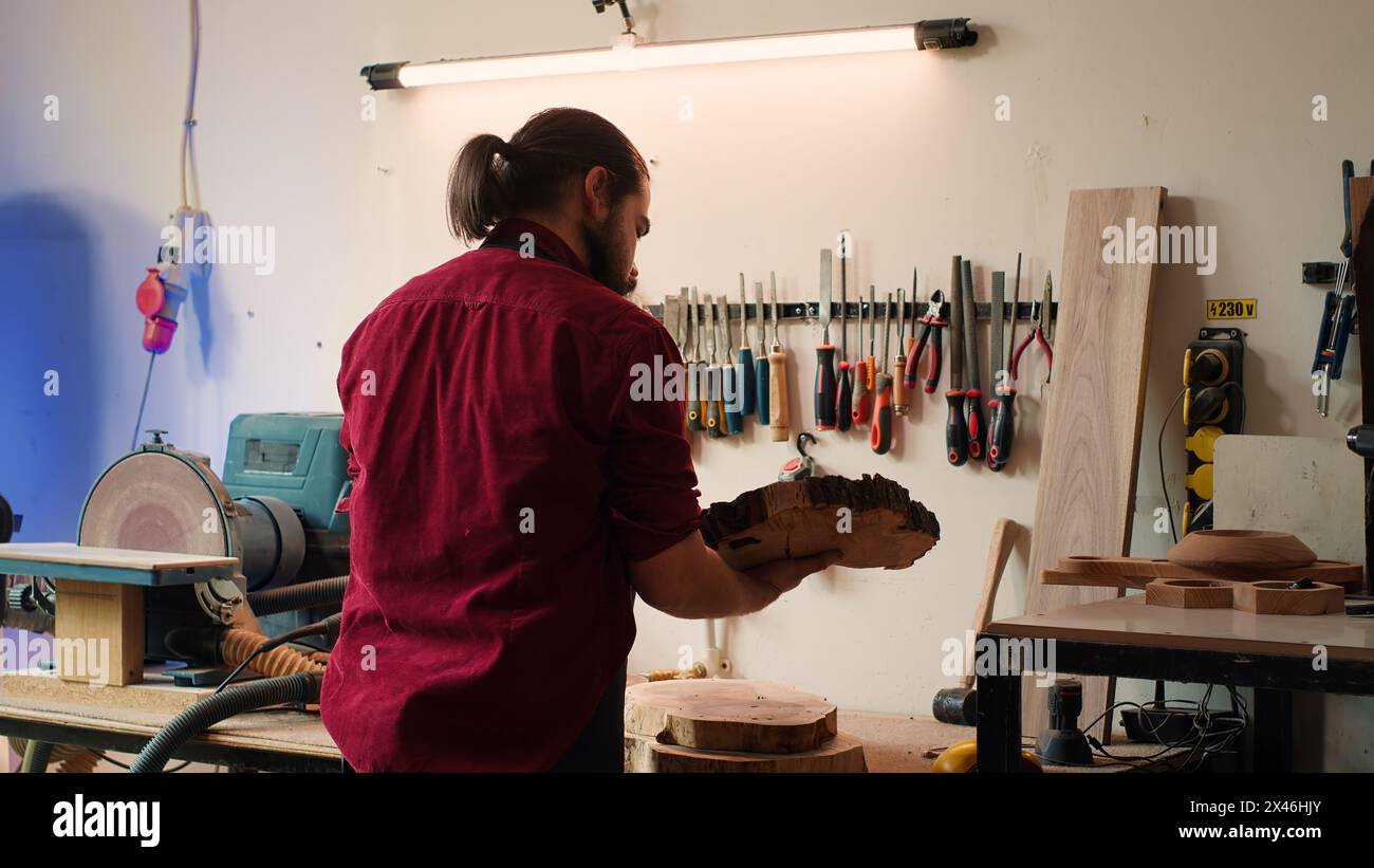 Zimmermann, der das Werkzeug vom Gestell zum Einsatz auf Holzmaterialien auswählt, unterstützt von einem afroamerikanischen Kollegen. Handwerker, der Qualitätssicherung an Holzstapel mit BIPOC-Lehrling, Kamera A, macht Stockfoto