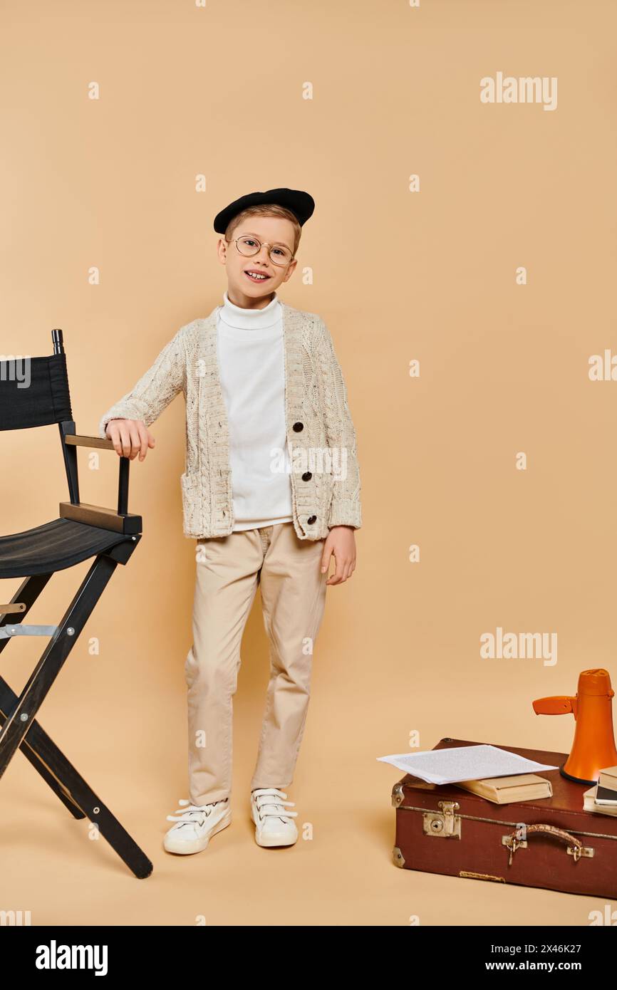 Ein niedlicher Junge, der als Filmregisseur gekleidet ist, steht neben einem Stuhl vor beigefarbenem Hintergrund. Stockfoto