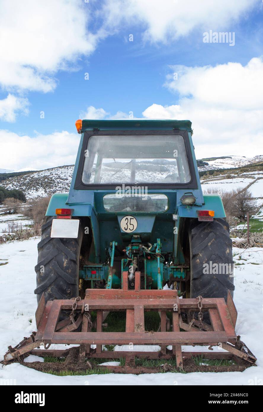 Alter Traktor mit Anbaugerät, das mitten auf einer schneebedeckten Wiese geparkt ist. Berglandwirtschaftskonzept Stockfoto