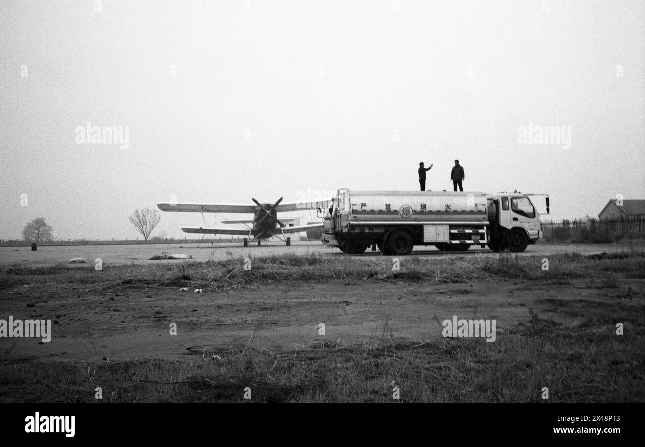 2013 stehen auf einem Landwirtschaftsflughafen in Dongying City, Provinz Shandong, China, zwei Personen auf einem Treibstoffwagen und bewegen sich in der Nähe eines stationären Propellerflugzeugs in einem offenen, scheinbar ländlichen Feld in Richtung Himmel. Das Foto fängt einen Moment der Interaktion zwischen den Menschen und der riesigen, leeren Umgebung ein, die sie umgibt, unter einem bewölkten Himmel. Stockfoto