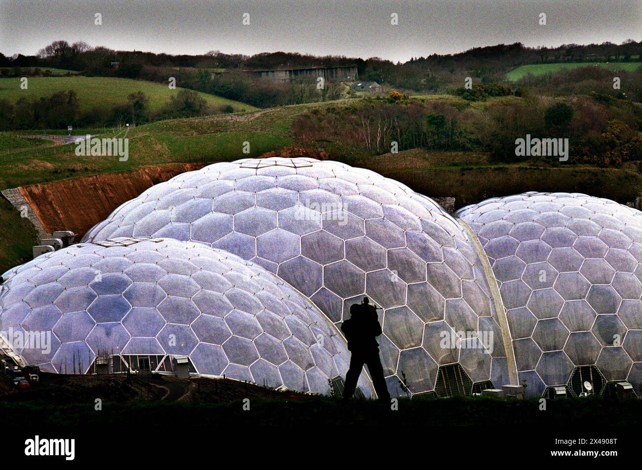 Eden Project Cornwall Mai 2001 das Eden Project (Cornish: Edenva) ist eine Besucherattraktion in Cornwall, England. Das Projekt befindet sich in einer zurückgewonnenen china-Tongrube, 2 km (1,2 mi) von der Stadt St Blazey und 5 km (3 mi) entfernt. der Komplex wird von zwei riesigen Gehegen dominiert, die aus benachbarten Kuppeln bestehen, die Tausende von Pflanzenarten beherbergen.[3] jedes Gehege emuliert ein natürliches Biom. Die Biome bestehen aus Hunderten sechseckiger und fünfeckiger Ethylentetrafluorethylen (ETFE)-Zellen, die von geodätischen Stahlrohrkuppeln unterstützt werden. Je größer Stockfoto