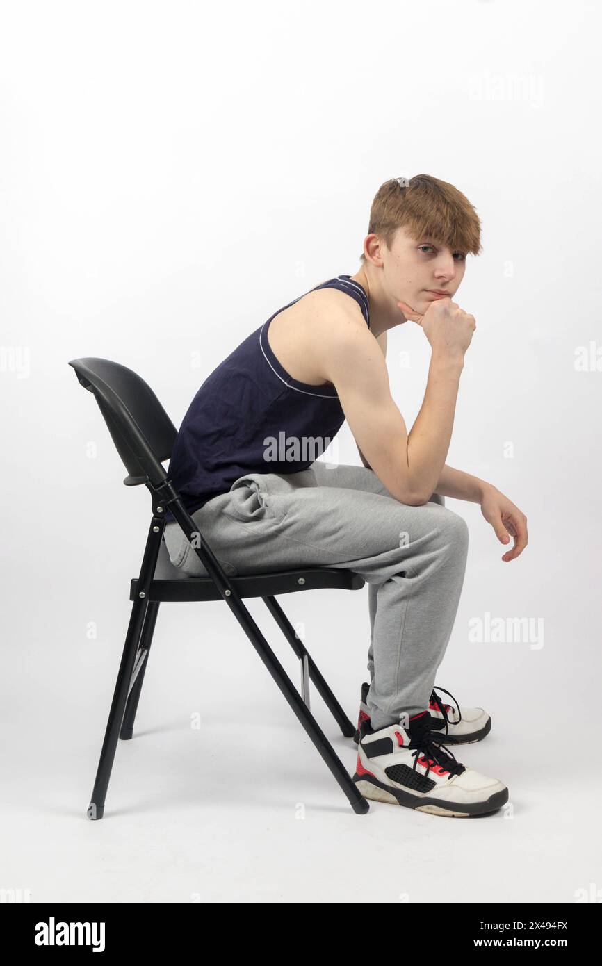 Ein 15-jähriger Teenager in ärmellosem Oberteil und Trainingshosen, der seitlich auf Einem Stuhl sitzt Stockfoto