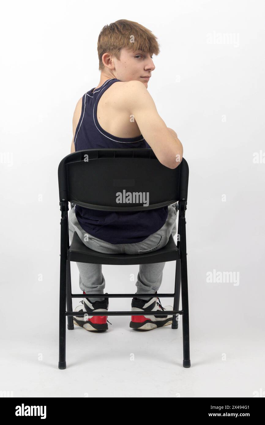 Ein 15-jähriger Teenager in ärmellosem Oberteil und Trainingshosen sitzt auf Einem Stuhl und blickt nach hinten Stockfoto