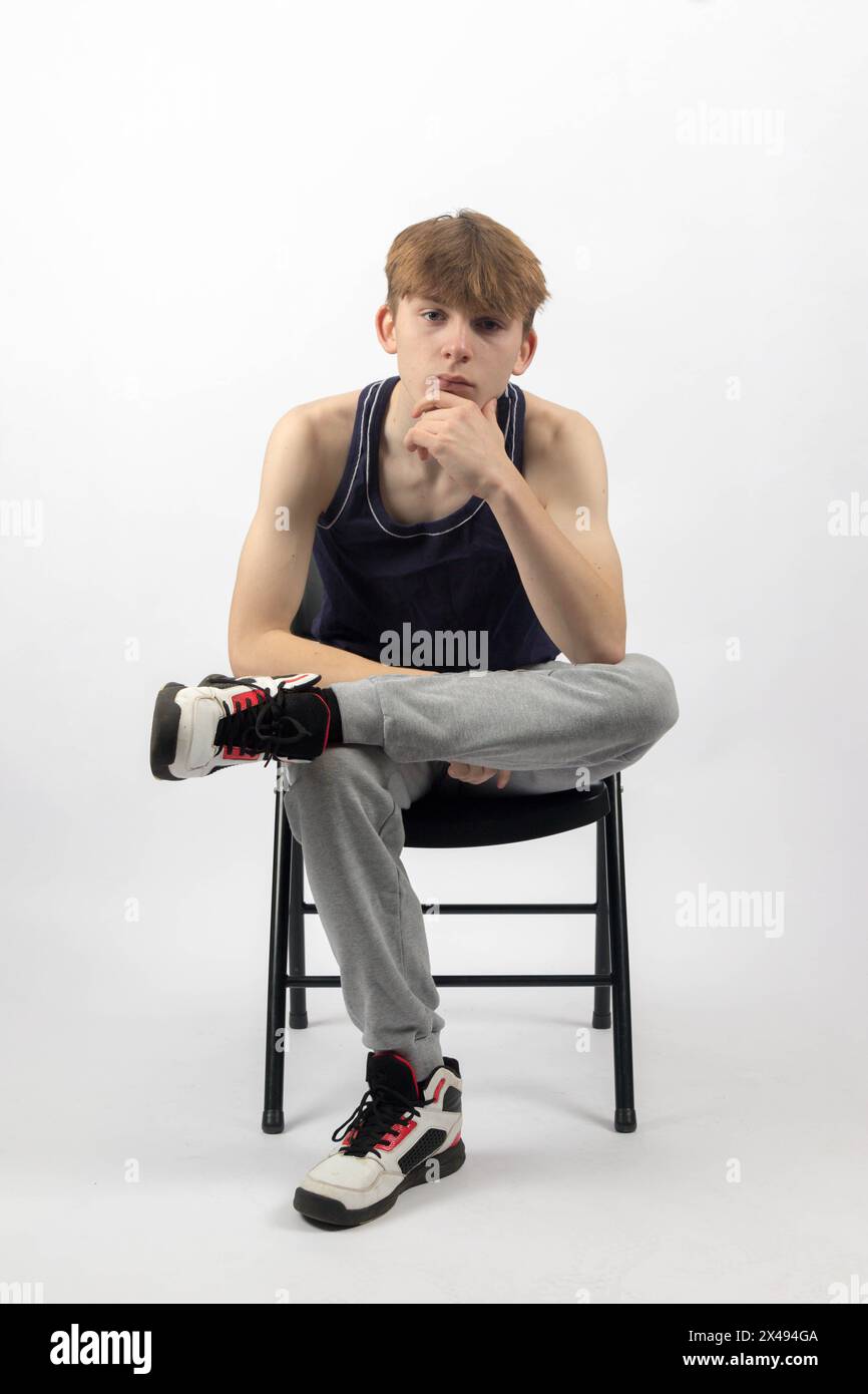 Ein 15-jähriger Teenager in ärmellosem Oberteil und Trainingshosen, der auf Einem Stuhl sitzt und sich nach vorne lehnt Stockfoto