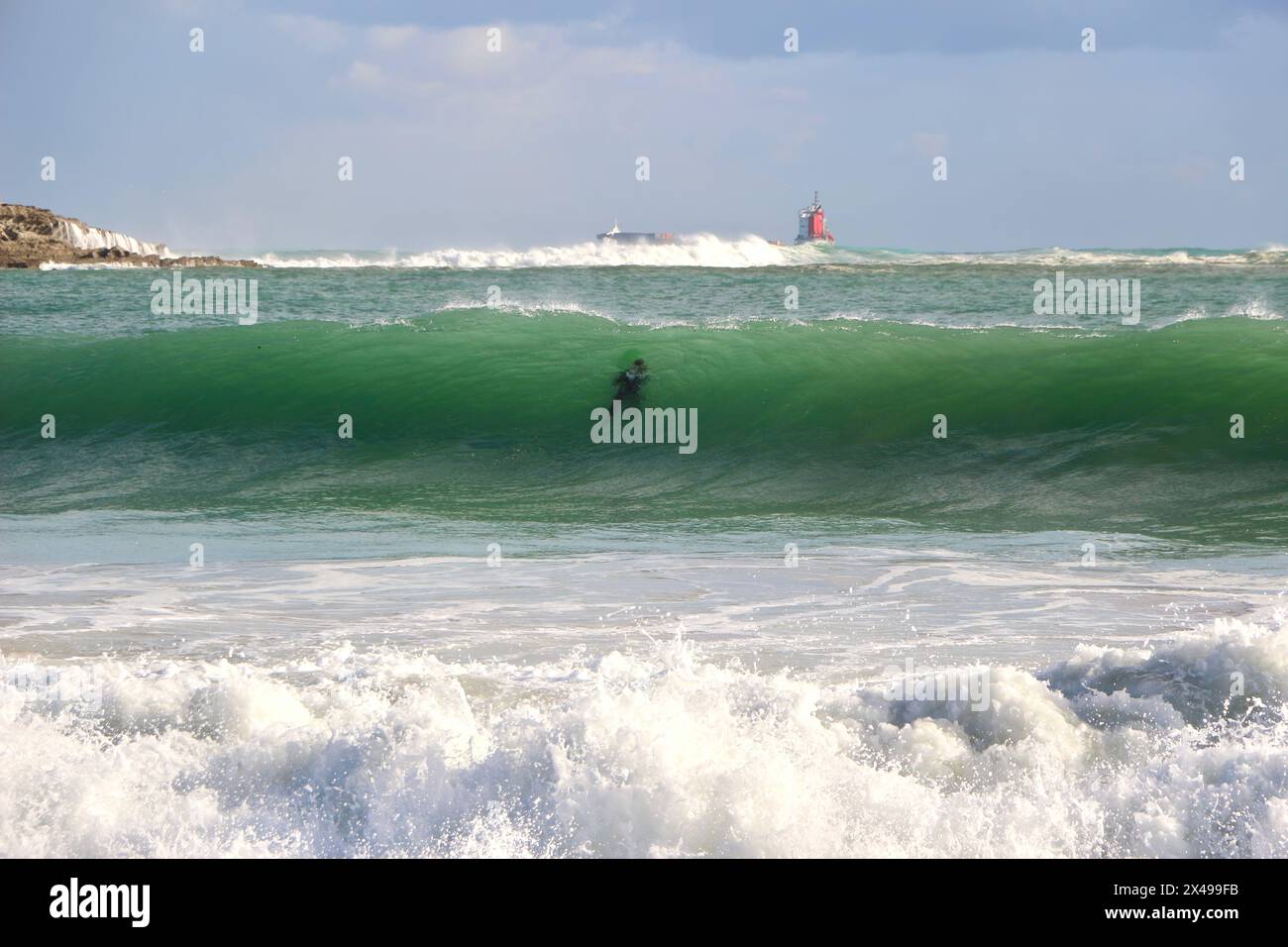 Untergetauchter Surfer in rauer See mit einem fernen Frachtschiff, das den Hafen von Santander an einem sonnigen Oktobermorgen verlässt Sardinero Santander Cantabria Spanien Stockfoto