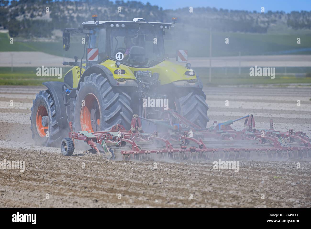 Ein Landwirt mit ihrem Traktor, der einen Pflug trägt und dabei große Felder mit Erntegut harkt und viel Staub aufwirbelt Stockfoto