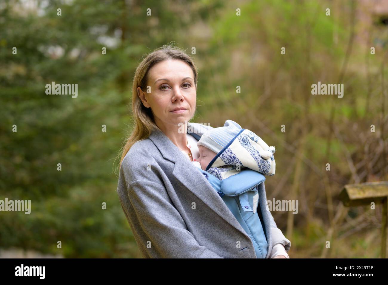 Oberkörperporträt einer glücklichen Frau, die in die Kamera schaut, während sie sie in einer Babytrage im Park hält und trägt Stockfoto