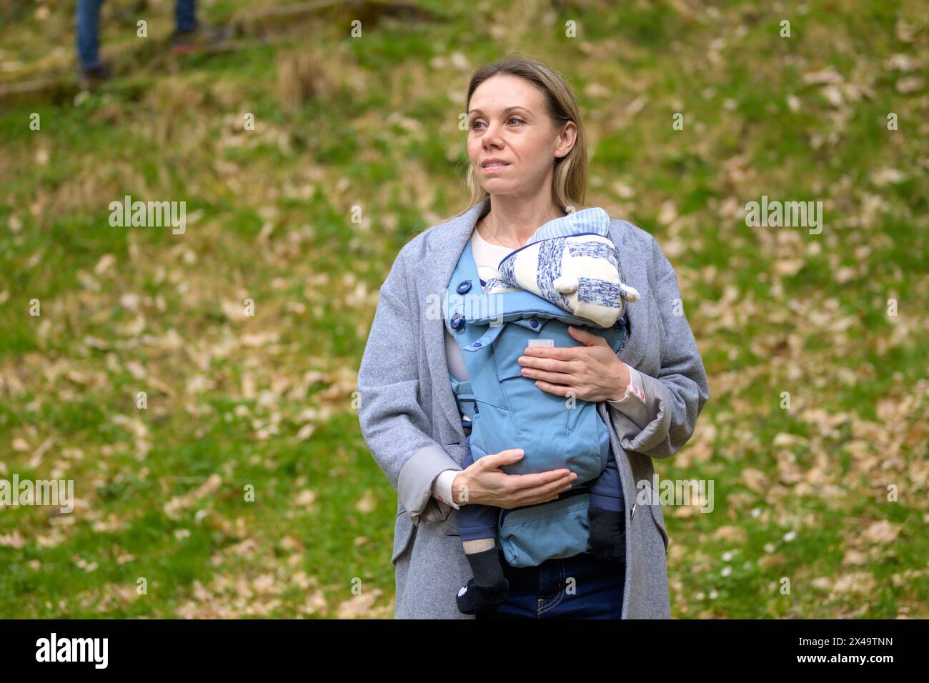 Glückliche Frau, die sich zur Seite schaut, während sie es in einer Babytrage im Park hält und trägt Stockfoto