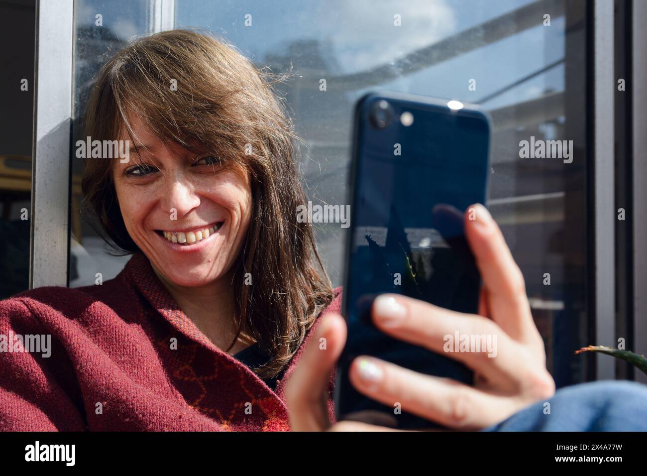 Lateinische Erwachsene Frau, sehr glücklich lächelnd, fühlt sich sehr gut und sitzt morgens auf dem Balkon, sonnt sich und macht Selfie mit ihrem Handy Stockfoto