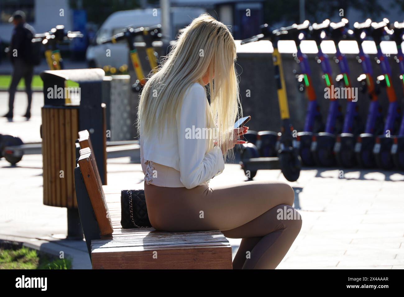 Schlankes Mädchen mit blonden, langen Haaren, das mit Smartphone an sonnigem Tag auf der Straßenbank von а sitzt Stockfoto