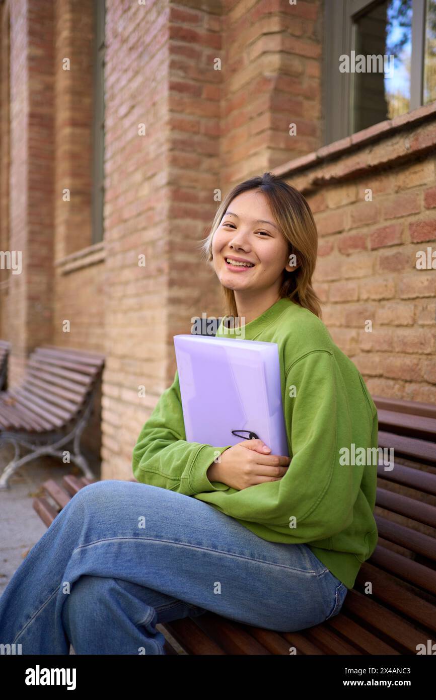 Porträt eines jungen, lächelnden chinesischen Studenten, der auf einer Bank auf dem Campus sitzt und in die Kamera schaut Stockfoto