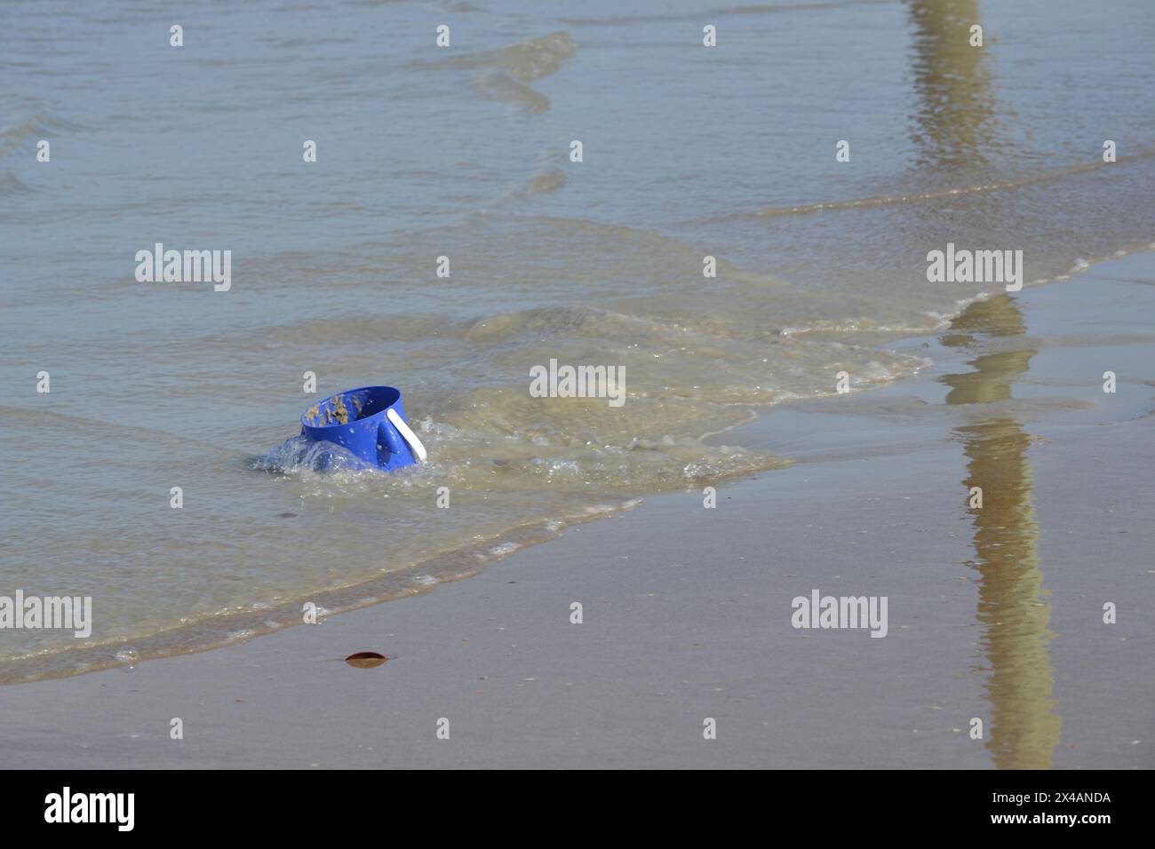 Ein verlassener blauer Eimer liegt zu drei Vierteln im Sand in der Nähe des Meeres, wo einfallende Wellen Reflexionen am Sandstrand erzeugen. Stockfoto
