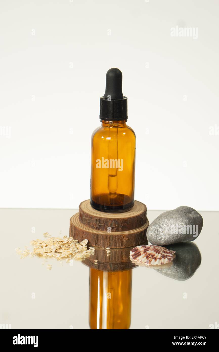 Eine braune Flasche ätherischen Öls auf einer glänzenden Oberfläche, die auf Holz sitzt und von Felsen, Muscheln und Hafer umgeben ist. Stockfoto