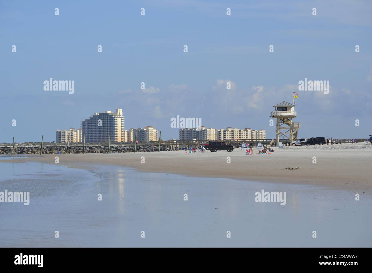 Vor der Kulisse des Ponce Inlet Strandes befinden sich eine Rettungswache, geparkte Autos und Menschen, die sich am Bootssteg mit der Skyline von New Smyrna Beach erholen. Stockfoto