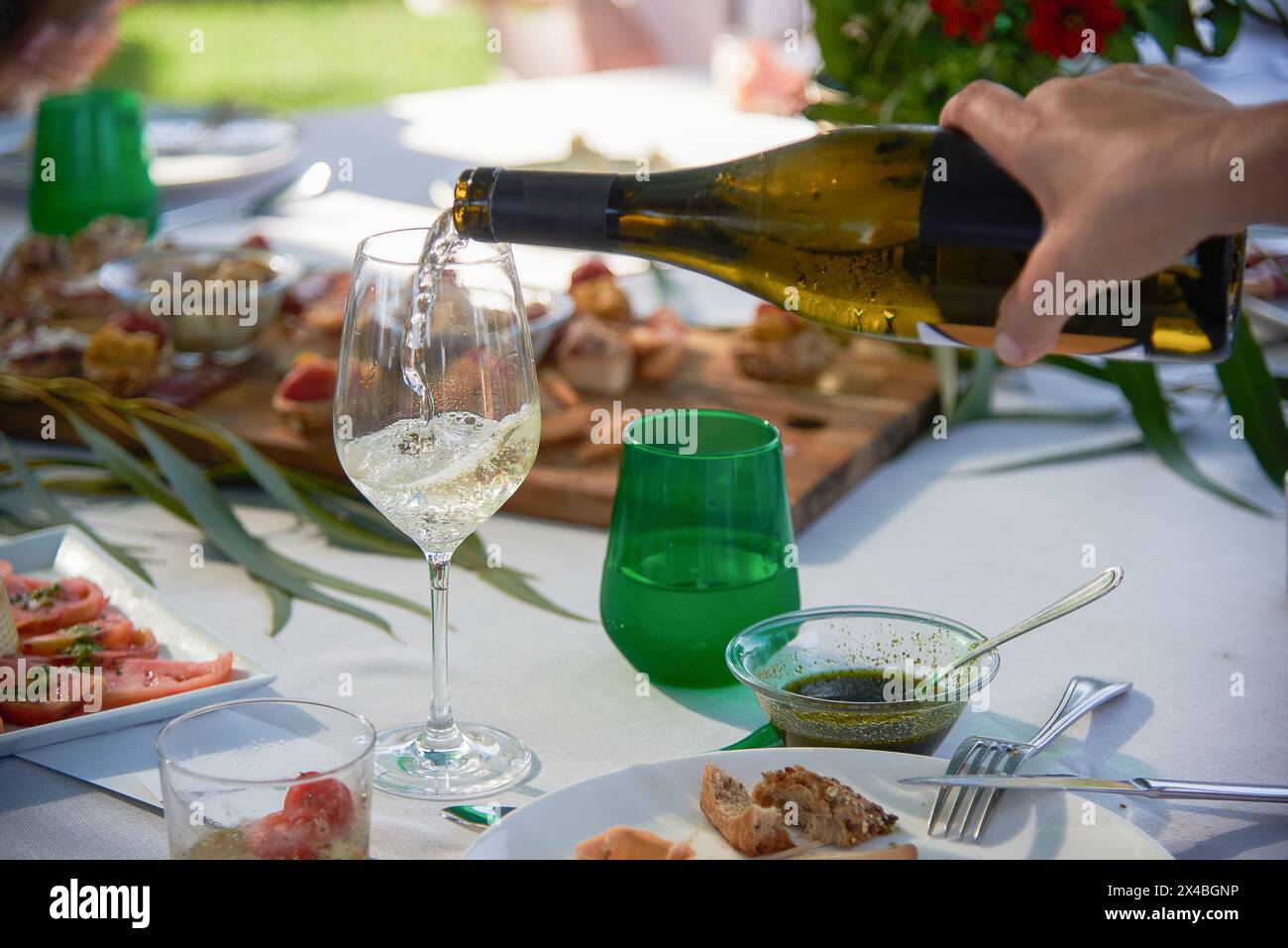 Geben Sie Weißwein von Hand in ein Glas, umgeben von köstlichen Vorspeisen. Festliche und sonnige Atmosphäre Stockfoto