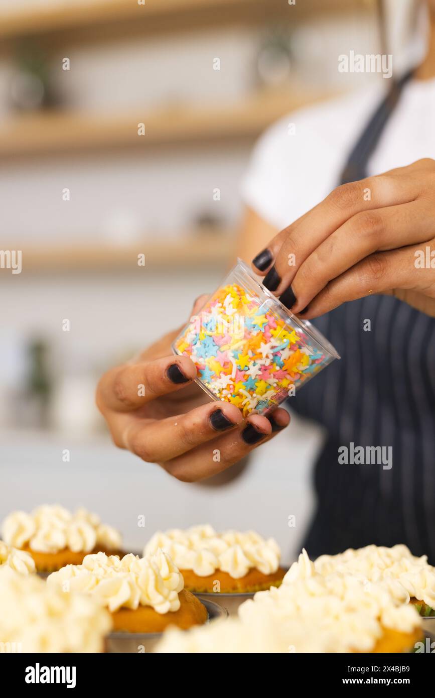 Eine reife asiatische Frau zu Hause, die Cupcakes mit bunten Streuseln dekoriert. Mit gestreifter Schürze, dunkel polierten Nägeln, mit intensiver Fokussierung o Stockfoto