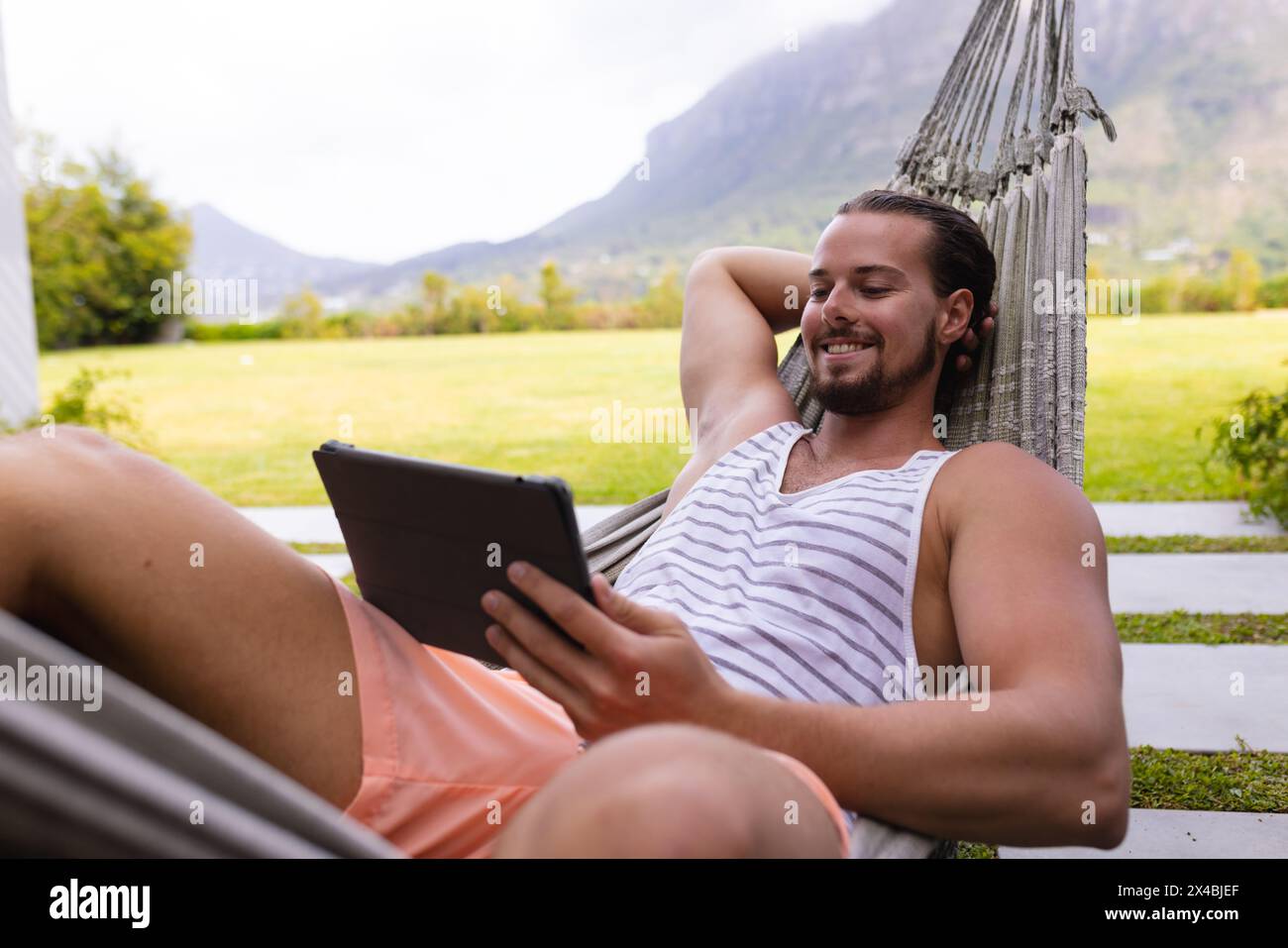 Bärtiger kaukasischer Mann entspannt sich in einer Hängematte mit einer Tablette. Er trägt legere Sommerkleidung und genießt einen friedlichen Moment in einem ruhigen blauen Hintergrund Stockfoto