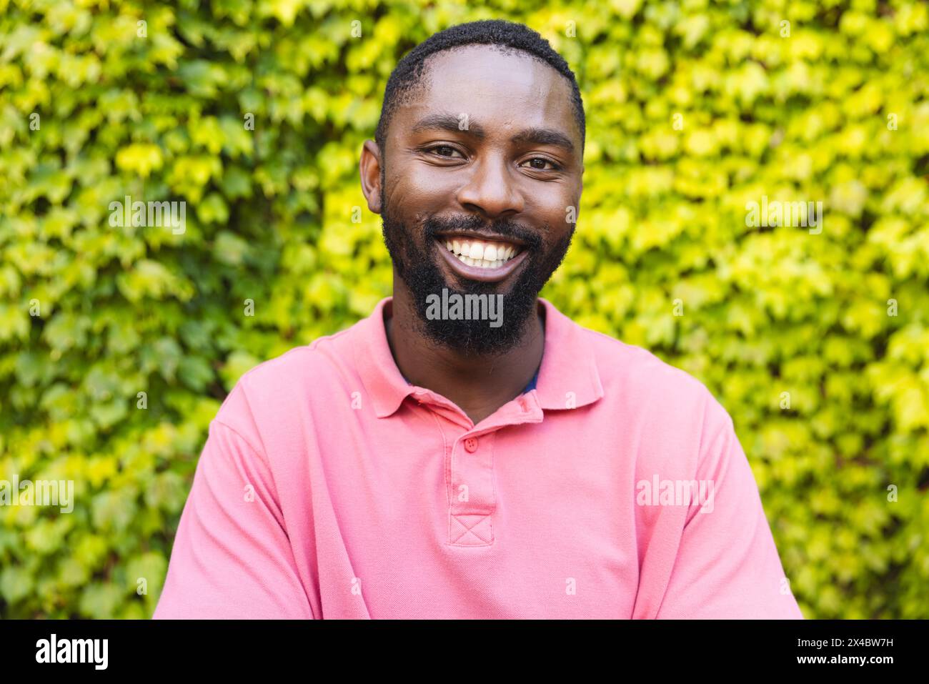 Ein Afroamerikaner in rosa Hemd, der draußen lächelt. Ein rosafarbenes Hemd, kurze schwarze Haare, einen Bart und helle Augen, frische Luft genießen, Stockfoto