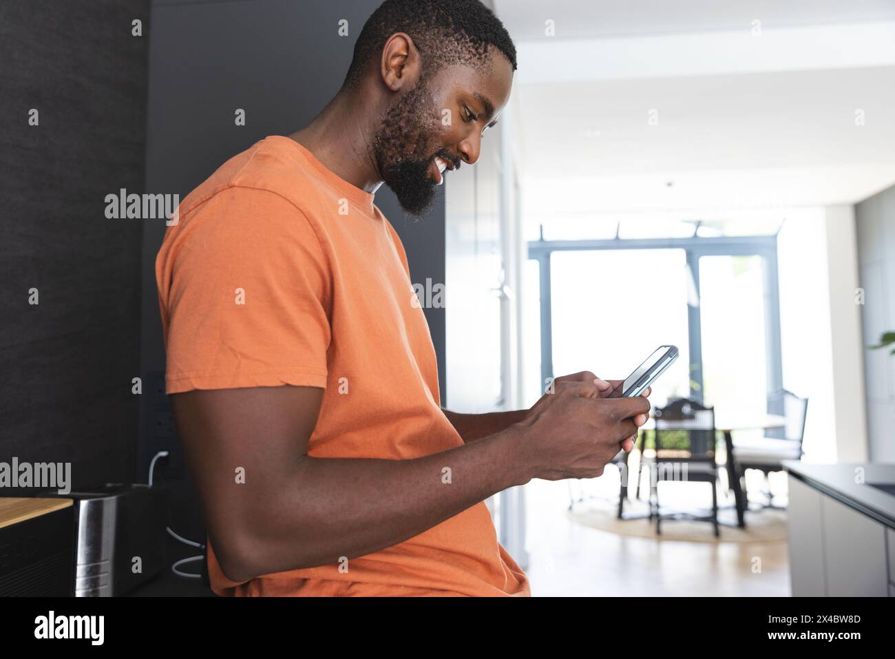 Zu Hause, Afroamerikaner, der in der Küche steht und auf das Smartphone blickt. Er trägt ein orangefarbenes Hemd, trägt kurze schwarze Haare und Bart und strahlt Casua aus Stockfoto