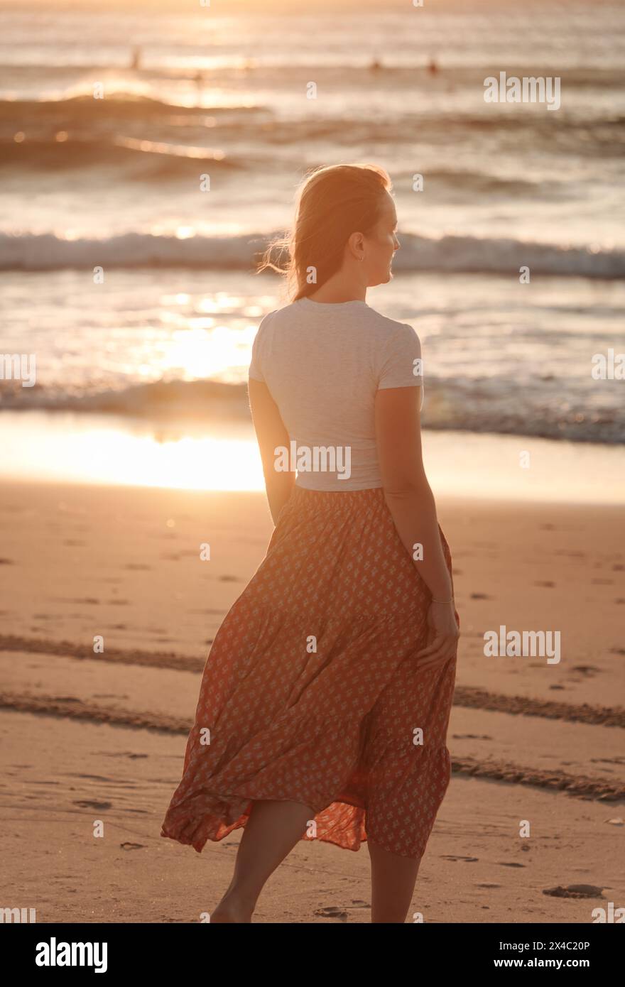 Eine schöne, einsame und nachdenkliche Frau am Meer, die in den Horizont blickt, am Strand im rötlichen Sonnenaufgang, mit ihrem Flowin Stockfoto