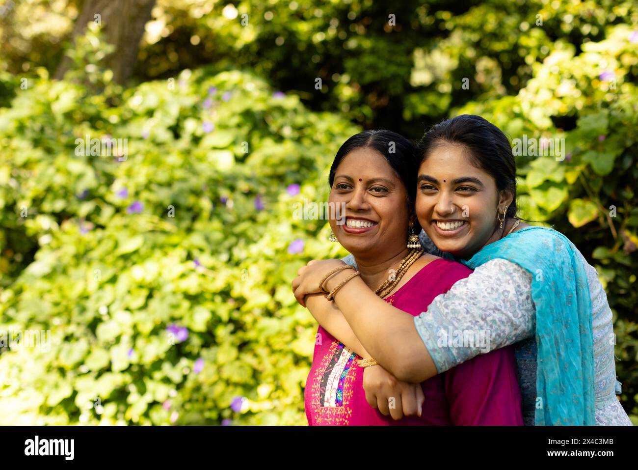 Indische Mutter und Teenager-Tochter umarmen, lächeln im Garten, Kopierraum. Beide haben dunkles Haar, mit Mutter kurz geschnitten und Tochter lang, unalt Stockfoto