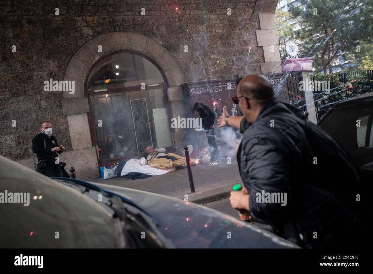 1. Mai 2024, Paris, Frankreich. An einem Maifeiertag zündeten Demonstranten ein Feuerwerk in der Nähe eines Schlafbereichs für Wohnungslose an, während ein anderer seine Wut über den Akt der Unachtsamkeit zum Ausdruck bringt. Eine schwarz gekleidete Person steht über dem Mann, um ihn zu beschützen. Quelle: Jay Kogler/Alamy Live News Stockfoto