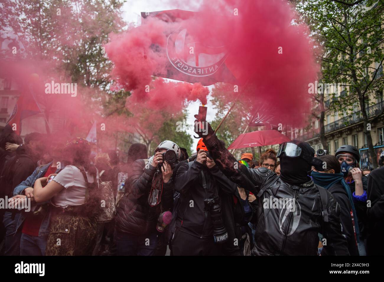 1. Mai 2024, Paris, Frankreich. Ein schwarz gekleideter Demonstrant am 1. Mai hält eine rote Rauchfackel vor einer Anti-Faschisten-Flagge. Quelle: Jay Kogler/Alamy Live News Stockfoto