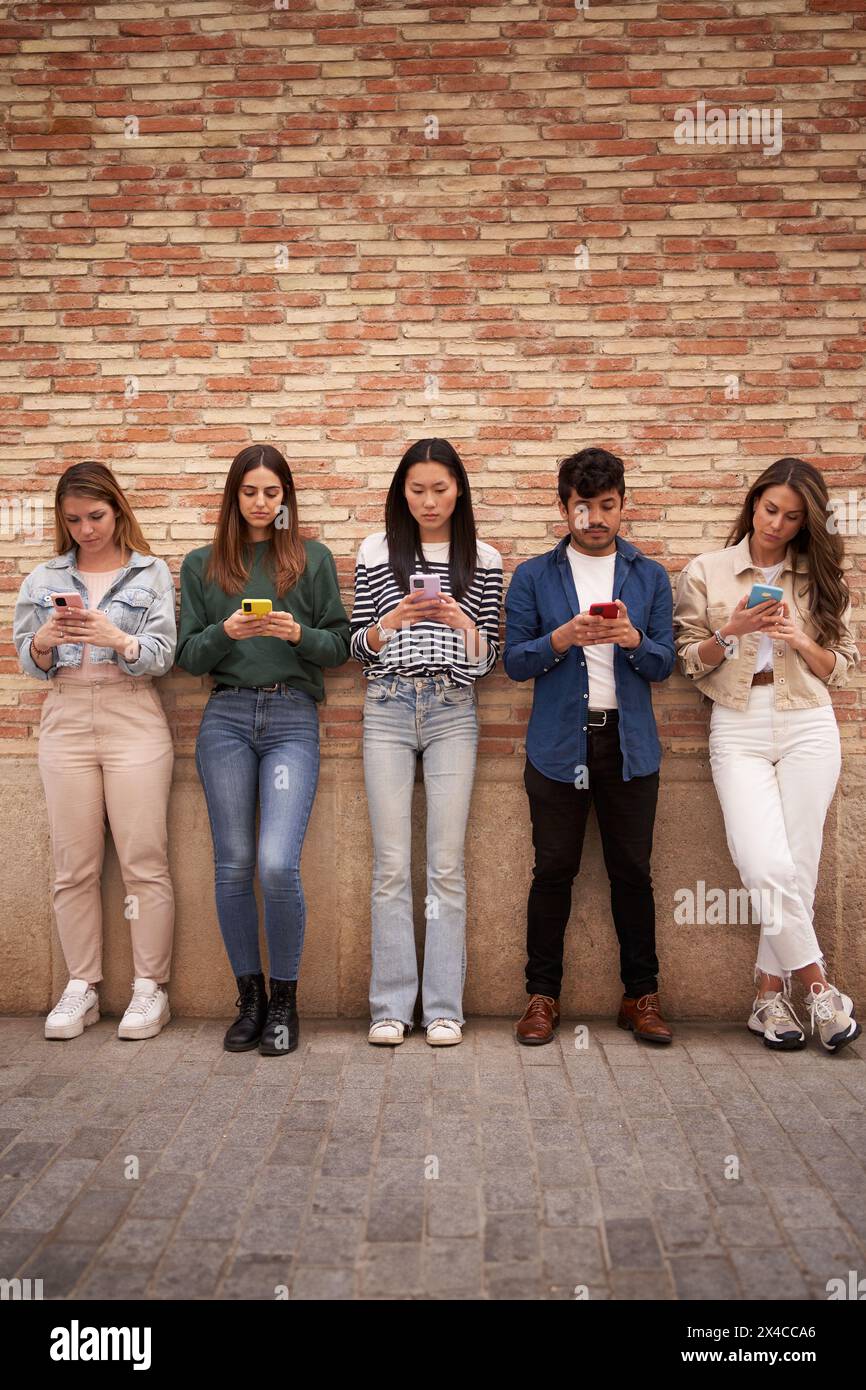 Multirassische ernsthafte Gruppe junger Generation Z, die sich mit Telefonen im Freien an einer Ziegelmauer stützt. Stockfoto