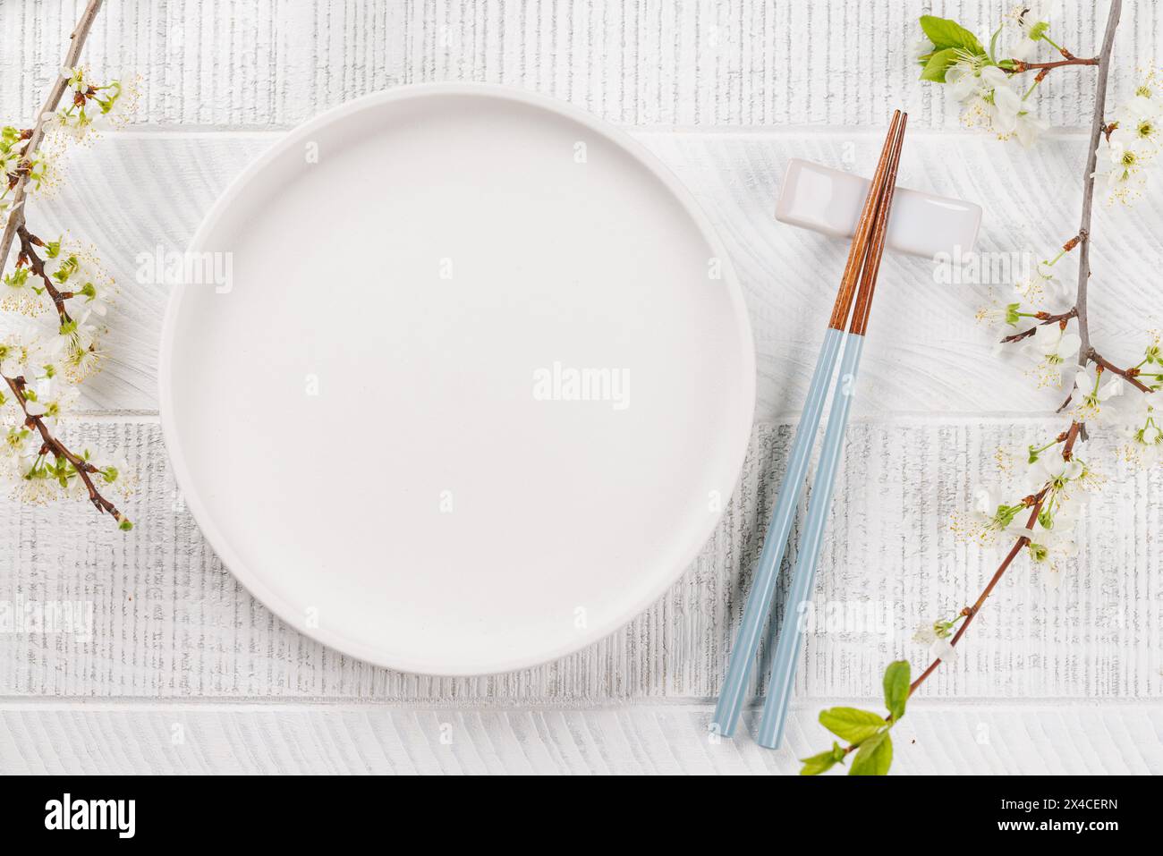 Ein leerer Teller auf dem Tisch, der mit Kirschblütenzweigen und Essstäbchen geschmückt ist, verkörpert die japanische Esskultur und bietet ausreichend Platz zum Kopieren Stockfoto