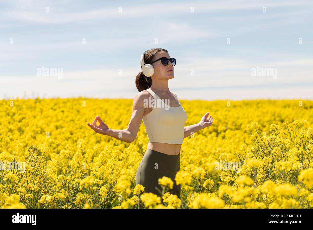 Sportliche Frau genießt Sonnenschein auf einem gelben Feld, meditiert und entkommt mit Kopfhörern. Stockfoto