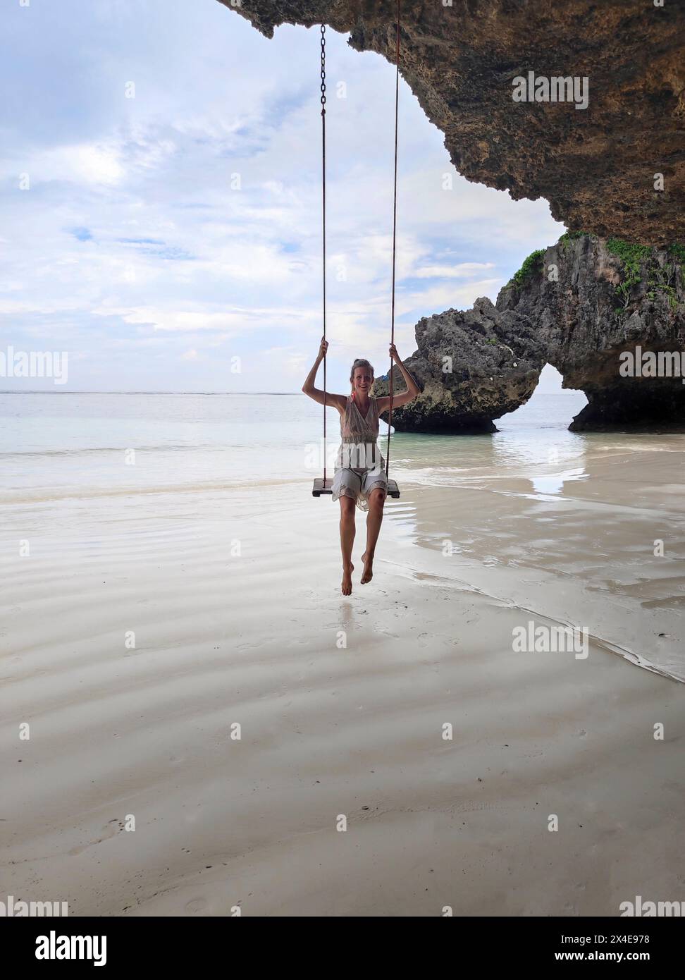 Eine junge Frau in einem Kleid, die das Leben auf einer Schaukel genießt, die an den Felsen hängt, am wunderschönen Strand von Mtende in Sansibar. Urlaub auf tropischer Insel. Stockfoto