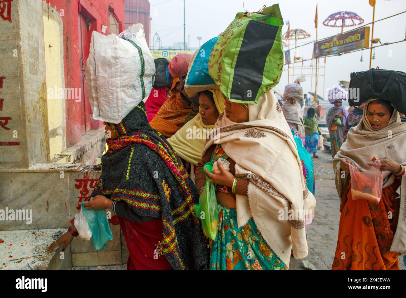 Pilgerinnen mit ausgeglichenen Taschen auf dem Kopf an einem kleinen Freiluftschrein in Dashwamedh Ghat in Varanasi, Indien. Stockfoto