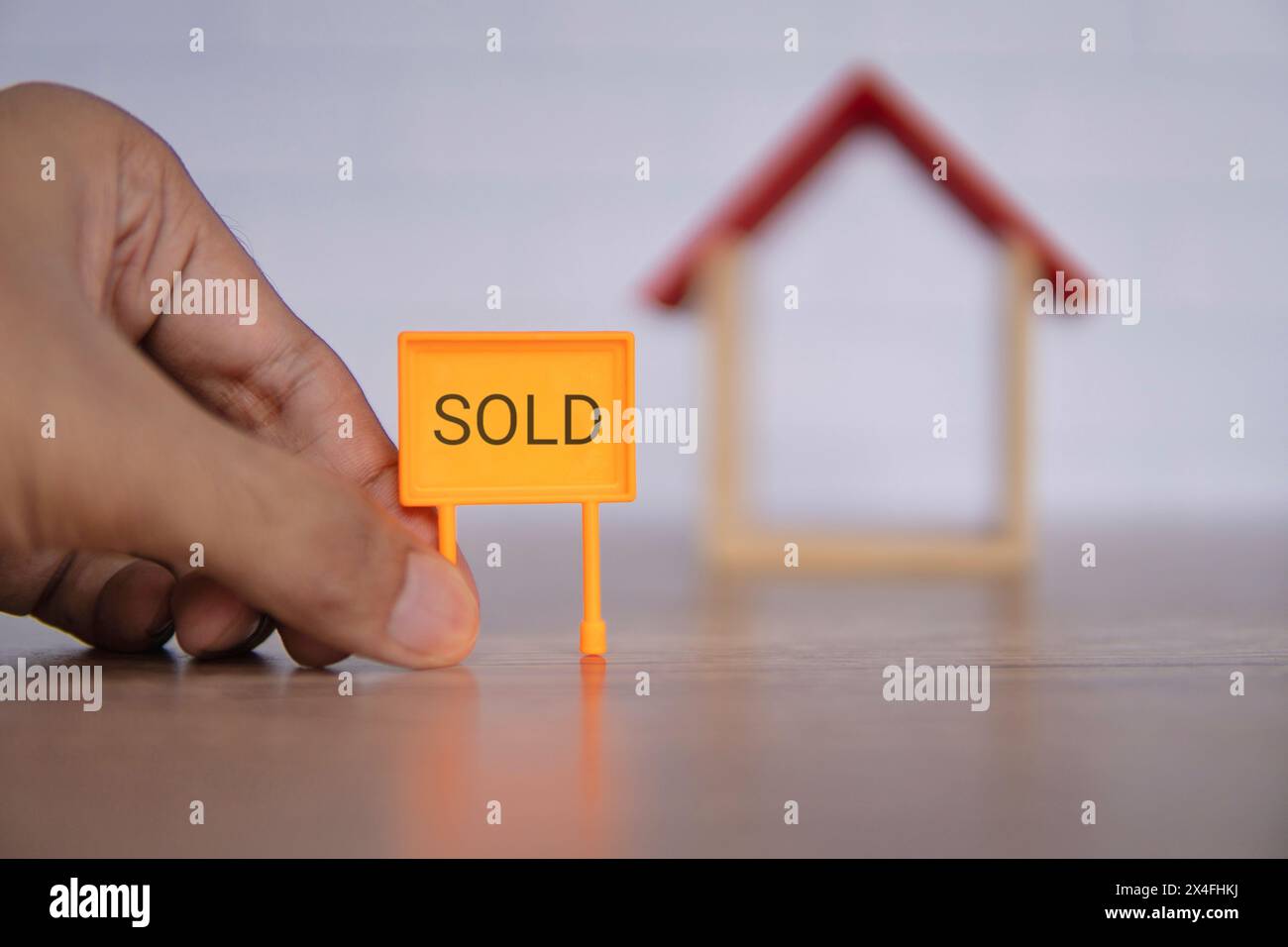 Ein Nahaufnahme-Foto einer Hand mit einem leuchtend orangefarbenen „VERKAUFT“-Schild vor einem hölzernen Miniaturhaus. Immobilien- und Immobilienkonzept. Stockfoto