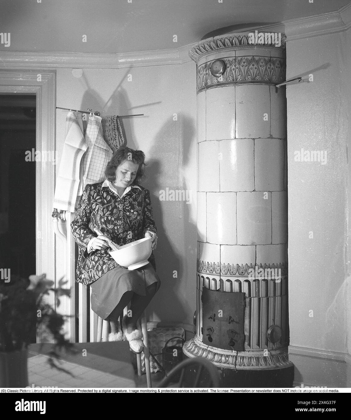 In den 1940er Jahren Das Innere eines Zimmers und eine junge Frau mit einer Schüssel im Knie, die den Inhalt quetscht. Sie sitzt neben einem Kachelofen, einer Holzheizquelle, die in der Vergangenheit üblich war. Die Wärme wurde lange Zeit in der großen Masse des Ofens gespeichert. Schweden 1946 Kristoffersson Ref. N138-3 Stockfoto