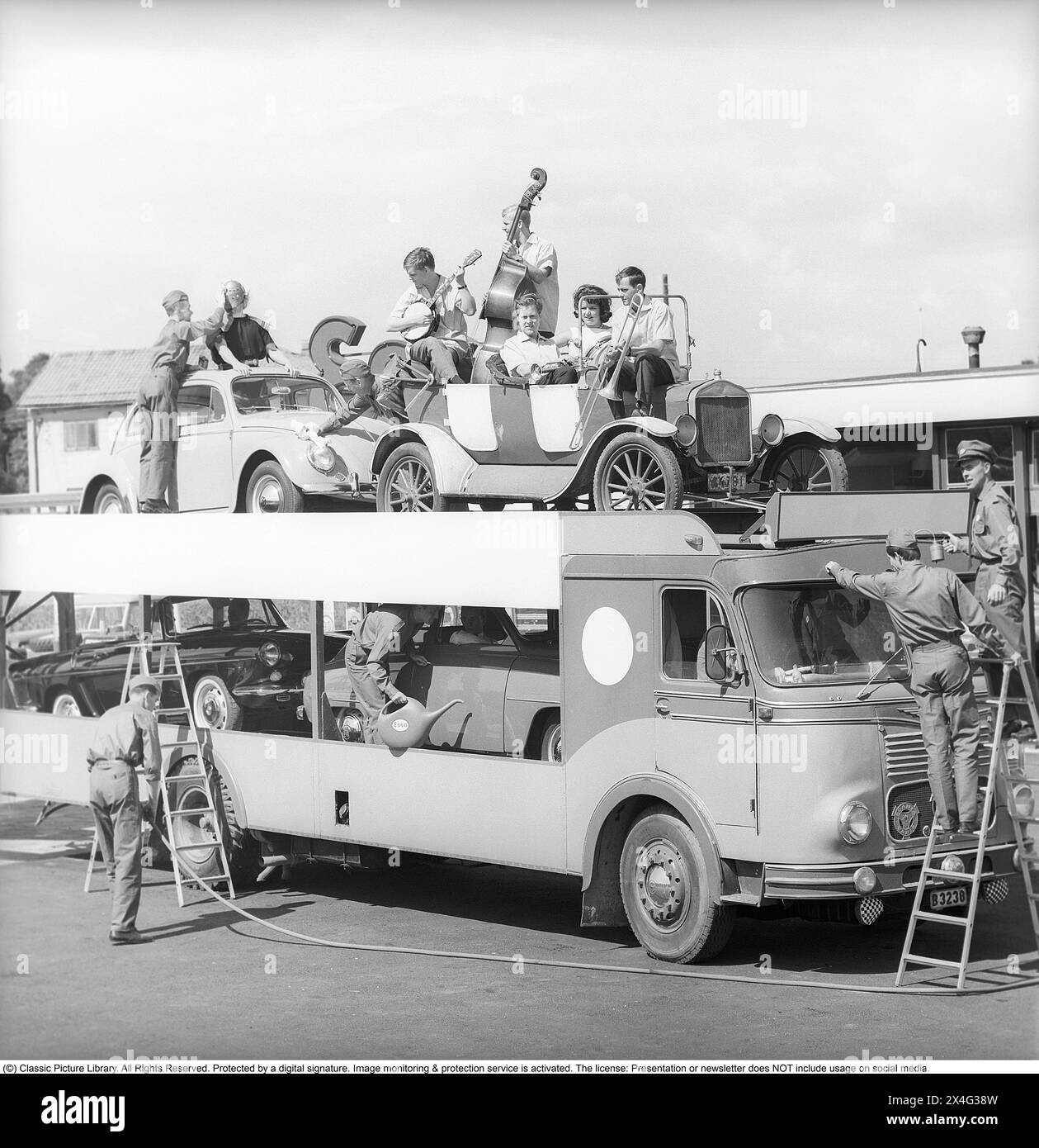Retro-Mitarbeiter an der Esso-Tankstelle. Ein lustiges Bild aus den 1960er Jahren, als eine Gruppe von Männern in der Kleidung des Unternehmens gesehen wird, die einem Lkw mit Autos verschiedener Modelle, einschließlich eines Veteranenautos mit einer Band mit ihren Instrumenten, dienen. Ein Bild, das während der Dreharbeiten eines Werbespots oder einer Werbung aufgenommen wurde. Die Firma Svenska Esso war eine Marke in Schweden zwischen den Jahren 1939 und 1987, als die Bahnhöfe ihren Namen in Statoil änderten und heute Circle K. Sweden 1963 trägt. Kristoffersson Ref. DC98-4 Stockfoto