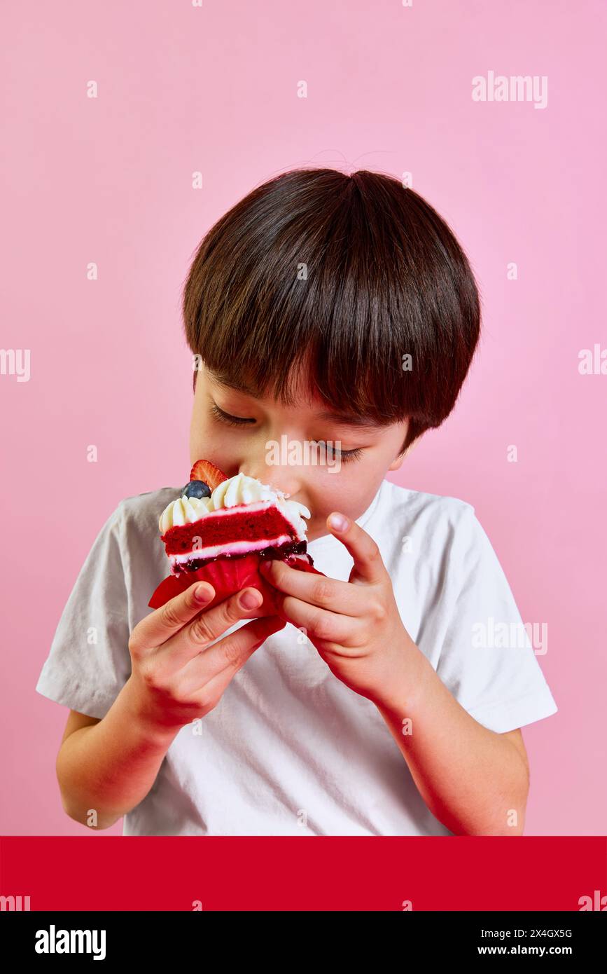 Herrliche Nachspeisezeit. Junge, Junge, der leckere rote Samtkuchen mit Sahne und Beeren genießt und vor rosa Hintergrund einen großen Biss macht Stockfoto