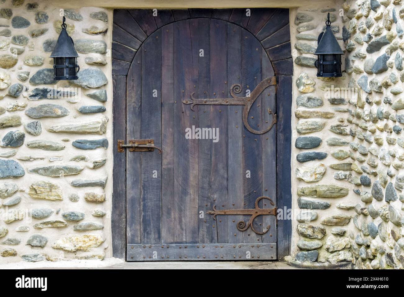 Die Steinmauer eines alten Hauses mit Lampen und einer Holztür mit geschmiedeten Elementen. Wunderschönes fabelhaftes Haus, märchenhaft. Kopierbereich. Selektiver Fokus Stockfoto Die Steinmauer eines alten Hauses mit Lampen und einer Holztür mit geschmiedeten Elementen. Wunderschönes fabelhaftes Haus, märchenhaft. Kopierbereich. Selektiver Fokus Stockfoto